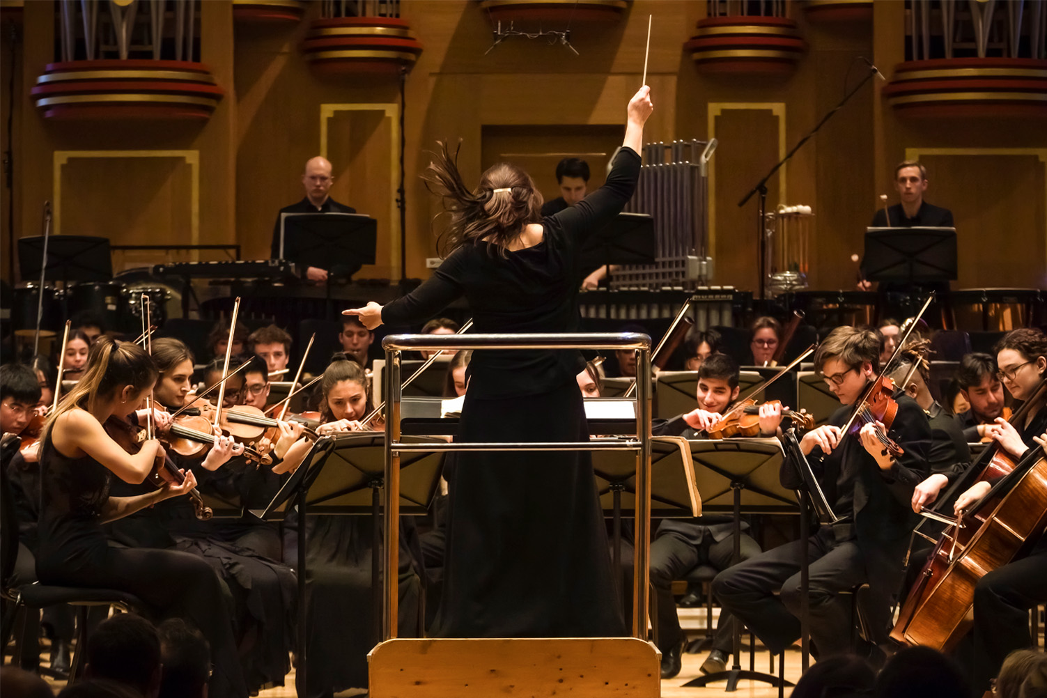 A women wearing a black dress, conducting an orchestra performance, with an organ in the background A women wearing a black dress, conducting an orchestra performance, with an organ in the background